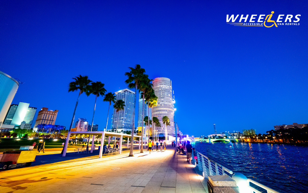 Evening view of Tampa Riverwalk with palm trees, waterfront lights, and modern buildings glowing under a deep blue sky. People walk and relax along the paved path beside the water.